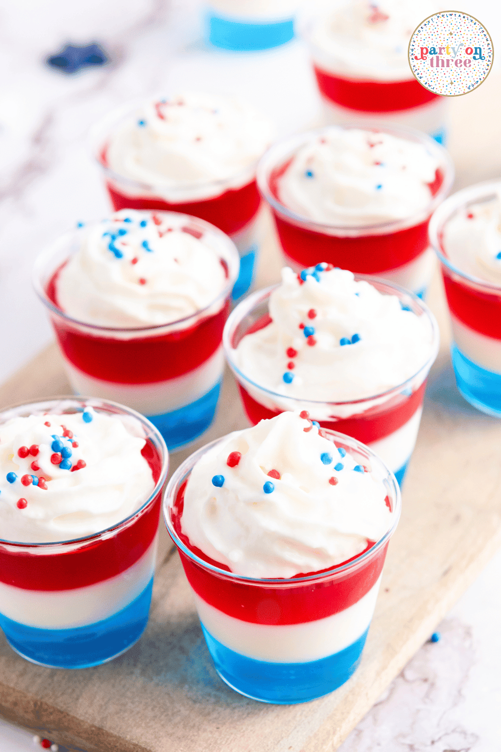 A tray of red, white, and blue layered Jello shots topped with whipped cream and patriotic sprinkles, perfect for a 4th of July party.
