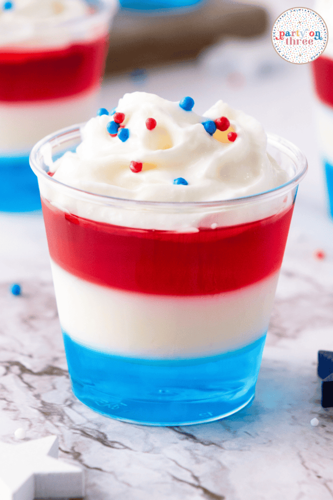 Close-up of a single layered Jello shot in red, white, and blue with whipped cream and red, white, and blue sprinkles on top.