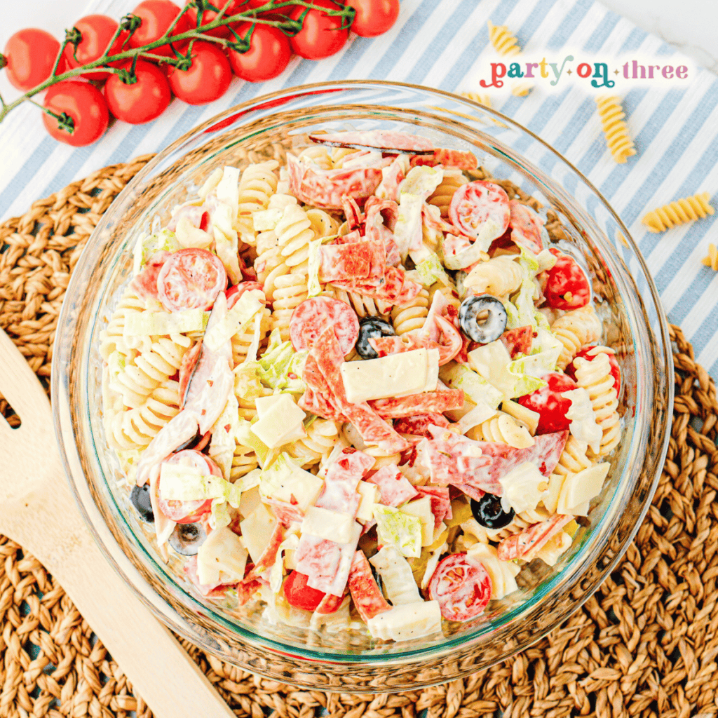 A large glass bowl of Italian grinder pasta salad next to tomatoes.