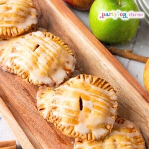 Close-up of glazed apple hand pies on a wooden tray beside a green apple and cinnamon sticks
