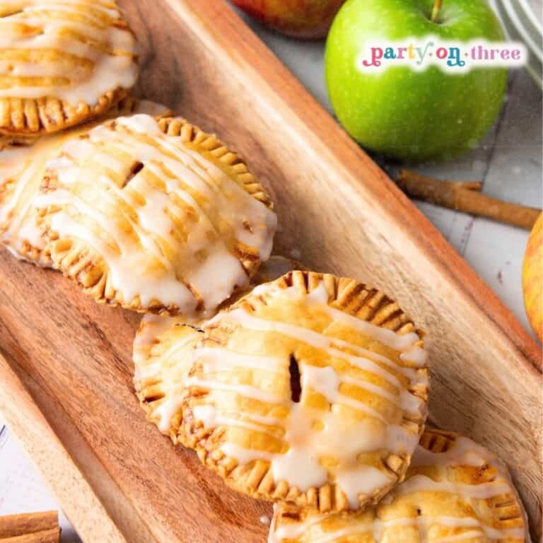 Close-up of glazed apple hand pies on a wooden tray beside a green apple and cinnamon sticks