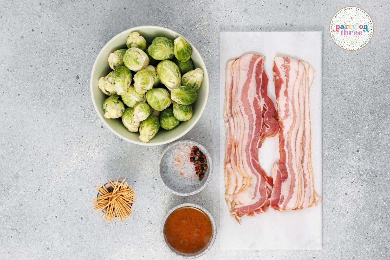 Overhead shot of raw brussels sprouts, bacon strips, toothpicks, salt, pepper and maple syrup