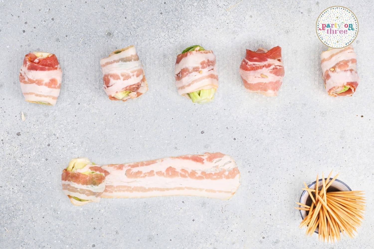 Bacon-wrapped brussels sprouts lined up unbaked next to a strip of bacon and a bowl of toothpicks