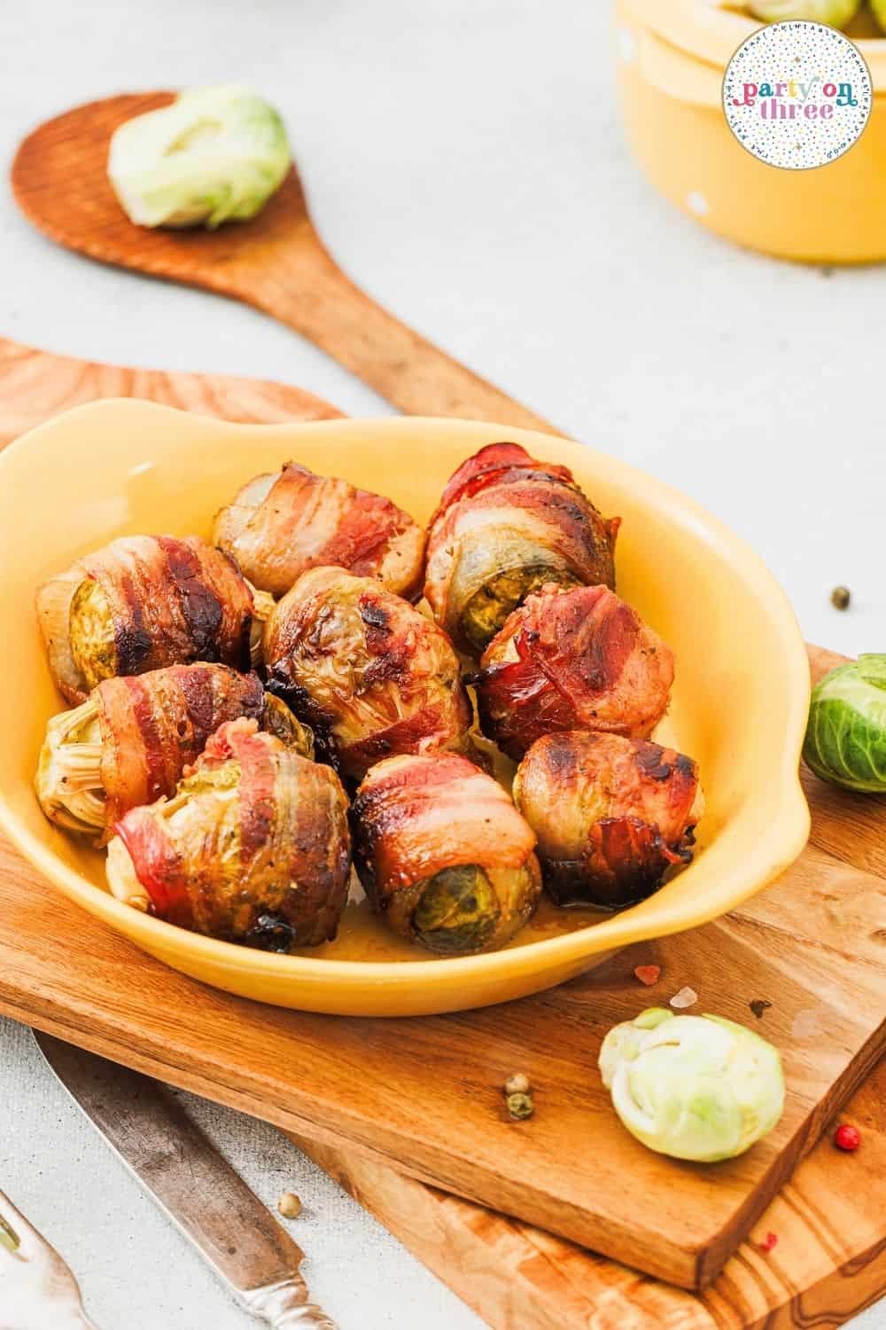 Baked bacon-wrapped brussels sprouts served in a yellow dish with wooden spoons nearby