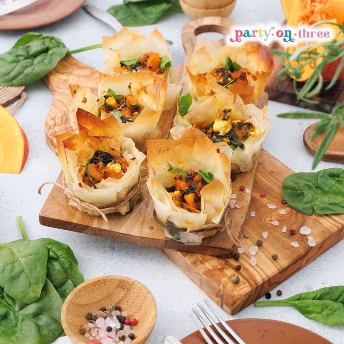 Baked pumpkin feta phyllo cups on a wooden board surrounded by spinach and pumpkin slices