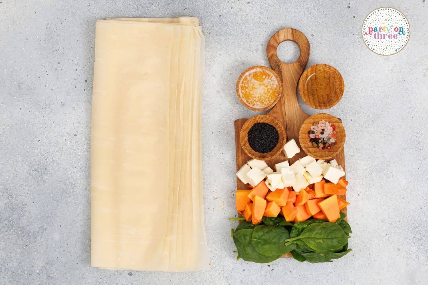 Phyllo dough sheets beside bowls of diced pumpkin, feta, spinach, and seasonings on a board