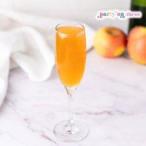 Apple Cider Mimosa in a champagne flute on a marble counter with apples and a bottle in the background