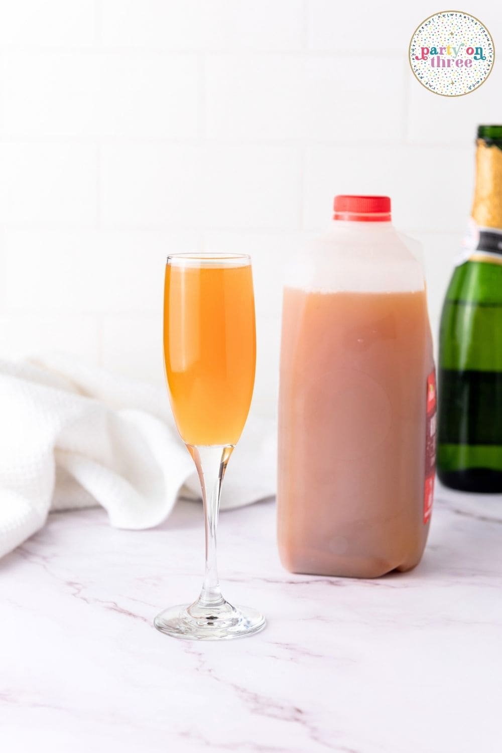 Champagne flute with orange liquid next to a jug of apple cider and a champagne bottle on a marble counter