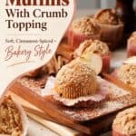 Apple cinnamon muffins with crumb topping on a wood board, with cinnamon sticks and apple slices nearby.