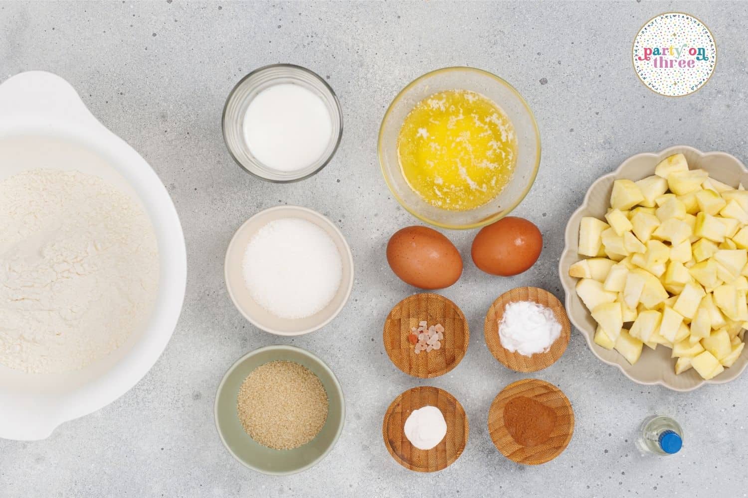 Ingredients for apple cinnamon muffins: flour, milk, melted butter, eggs, diced apples, sugar, and spices in bowls.