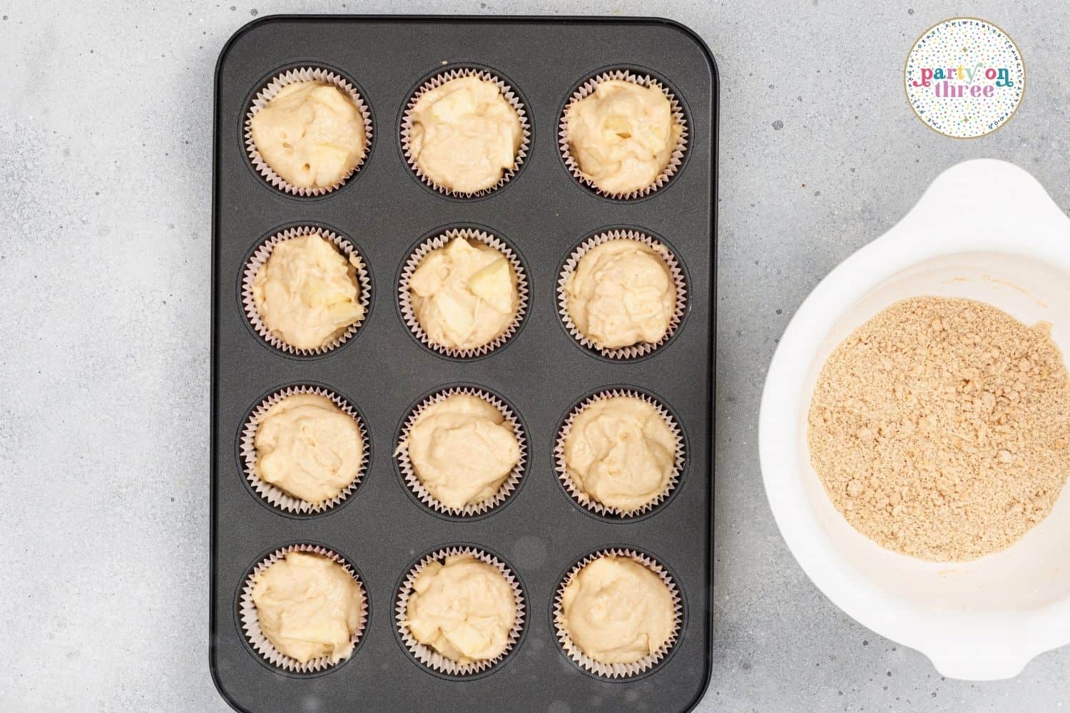 Muffin tin filled with apple cinnamon batter next to a bowl of crumble topping