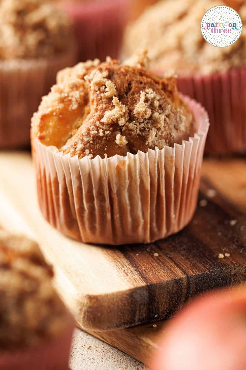 Baked muffin with crumb topping in paper liner, set on a wooden board with more muffins blurred behind.