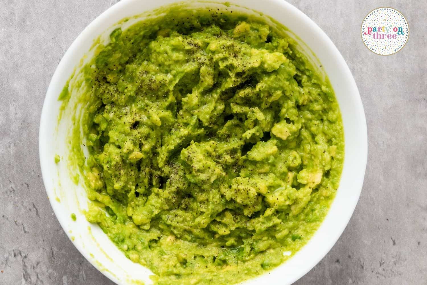 Mashed guacamole with cracked black pepper in a white bowl, ready for spreading on pita wedges.