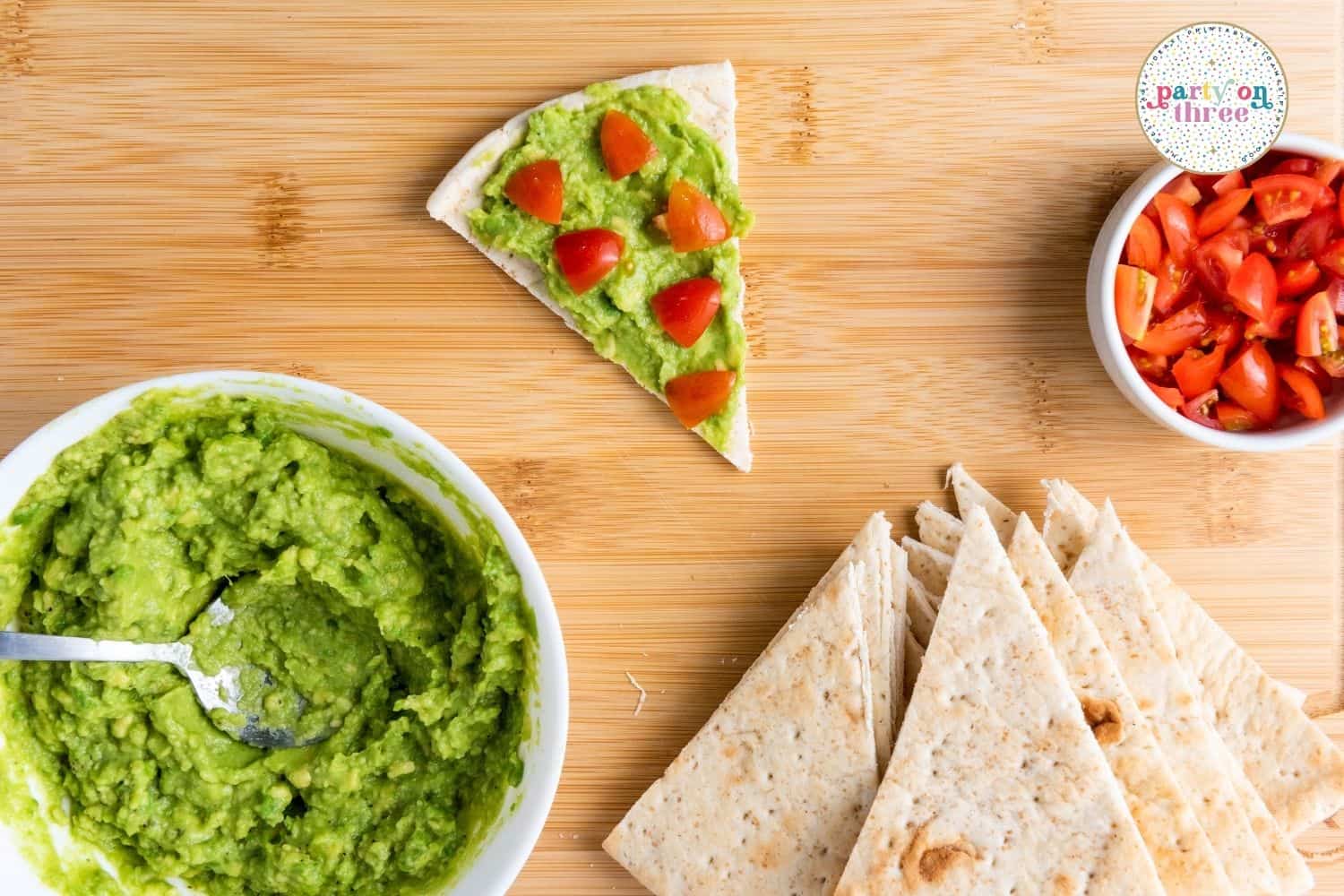 Mashed avocado in a bowl beside pita triangles, diced tomatoes, and a tomato-topped pita wedge.