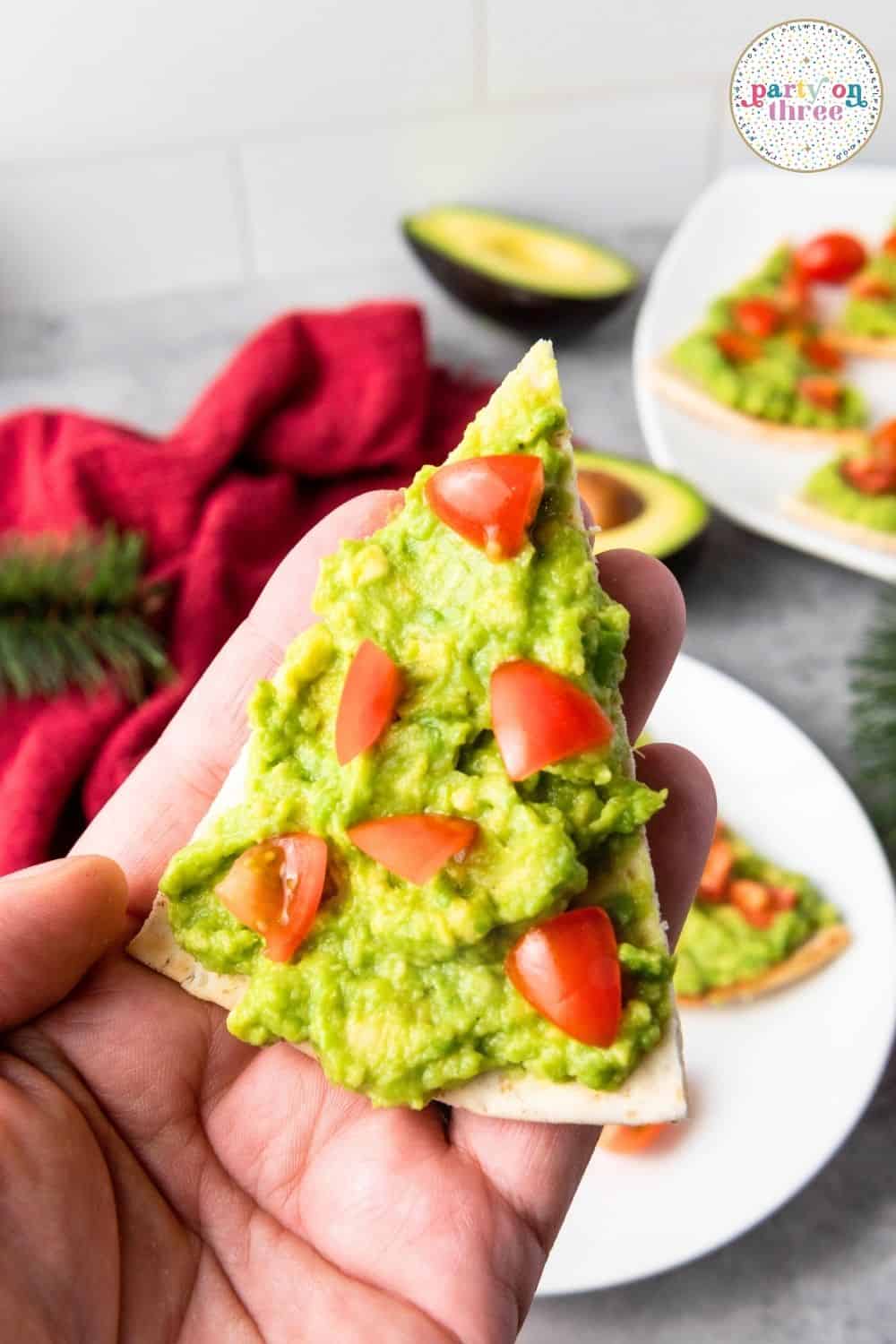 Hand holding a pita tree triangle topped with mashed avocado and diced tomato pieces as ornaments