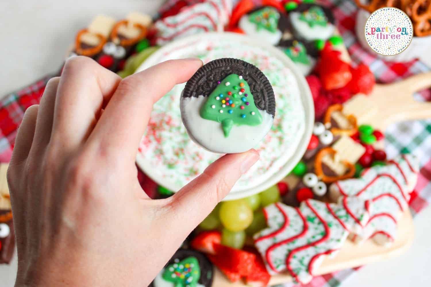 Hand holding a white chocolate-dipped Oreo decorated with a green Christmas tree, above a holiday dessert board.