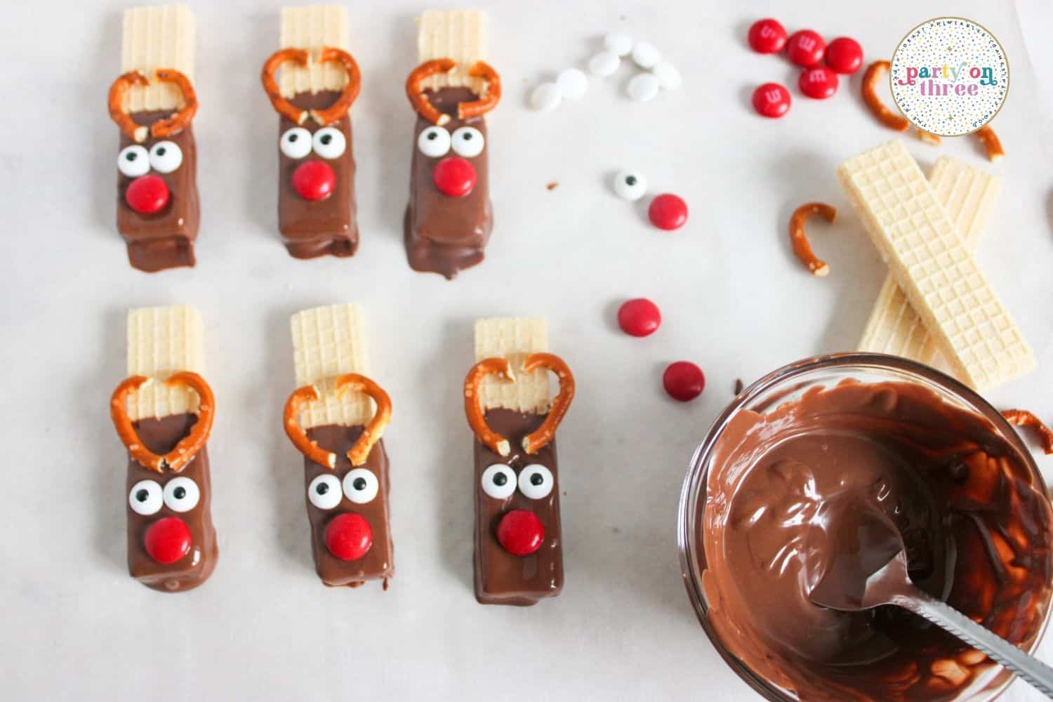 Chocolate-dipped wafer reindeer with pretzel antlers, candy eyes and red M&M noses on parchment paper
