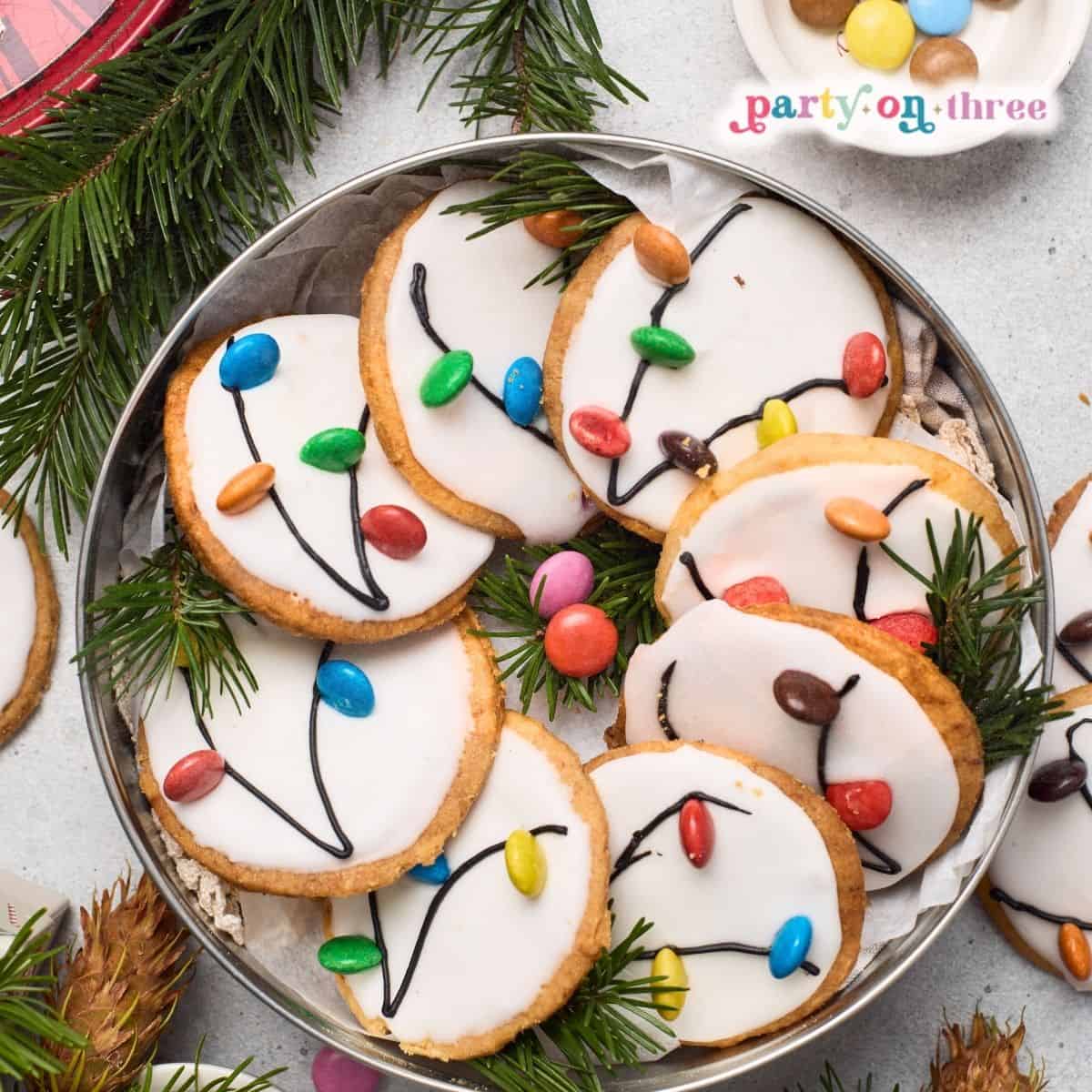 Christmas Light Cookies with white icing and candy "bulbs" piped with cord lines, arranged on a tin tray with greenery