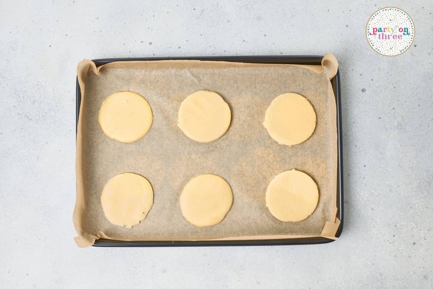 Six round sugar cookie dough rounds on a parchment-lined baking sheet before baking