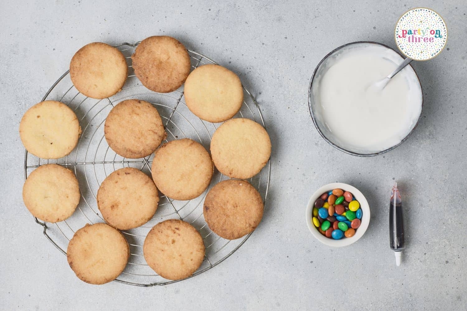 Round sugar cookies on a cooling rack beside bowls of white icing and colorful candies for decorating