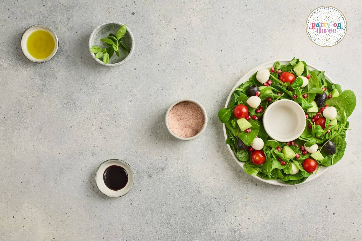 Wreath-shaped salad with greens, tomatoes, mozzarella, and olives next to small bowls of oil, herbs, salt, and balsamic.