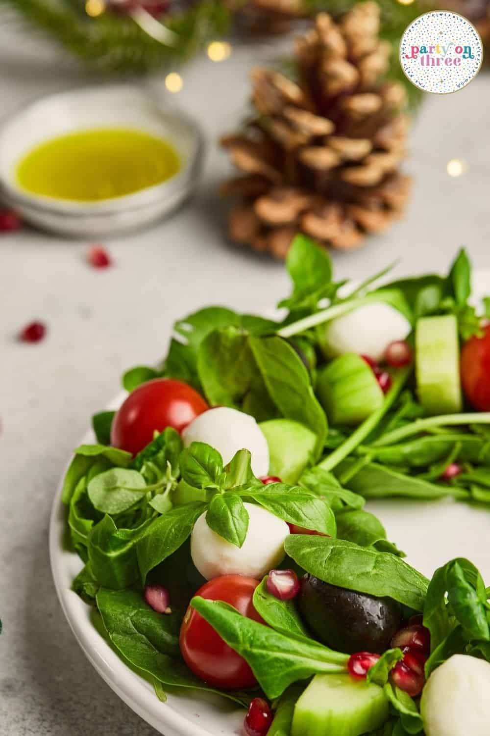 Wreath-shaped salad with basil, mozzarella, cherry tomatoes, olives, and pomegranate seeds on a white plate.