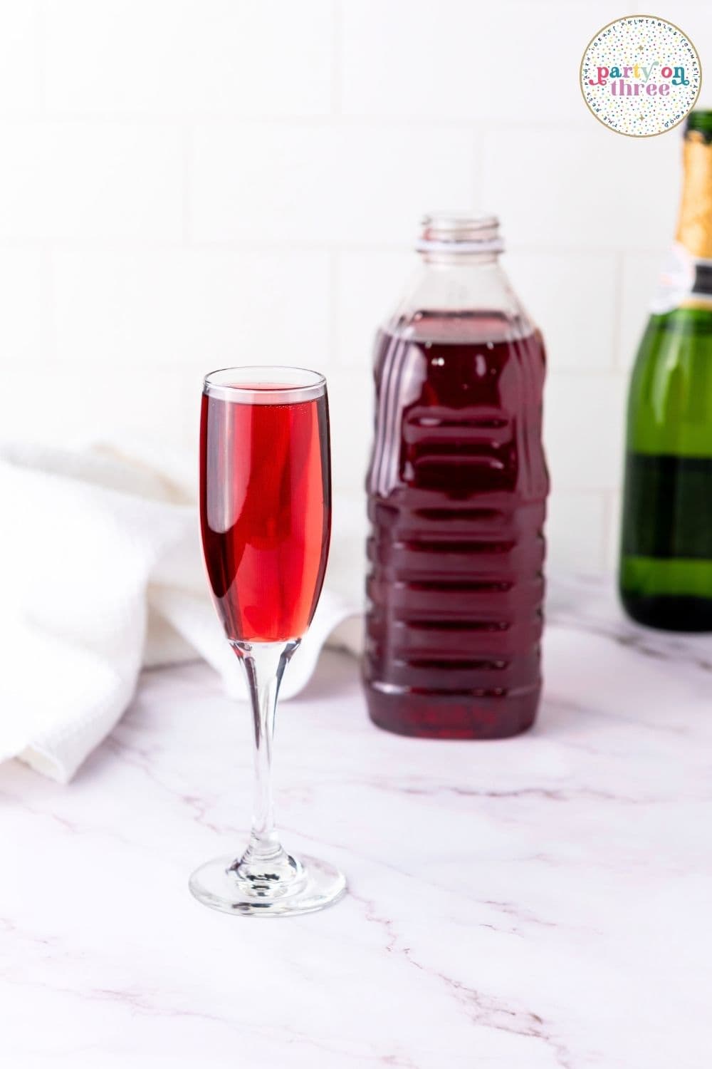 Champagne flute filled with cranberry mimosa beside cranberry juice bottle and champagne on marble counter