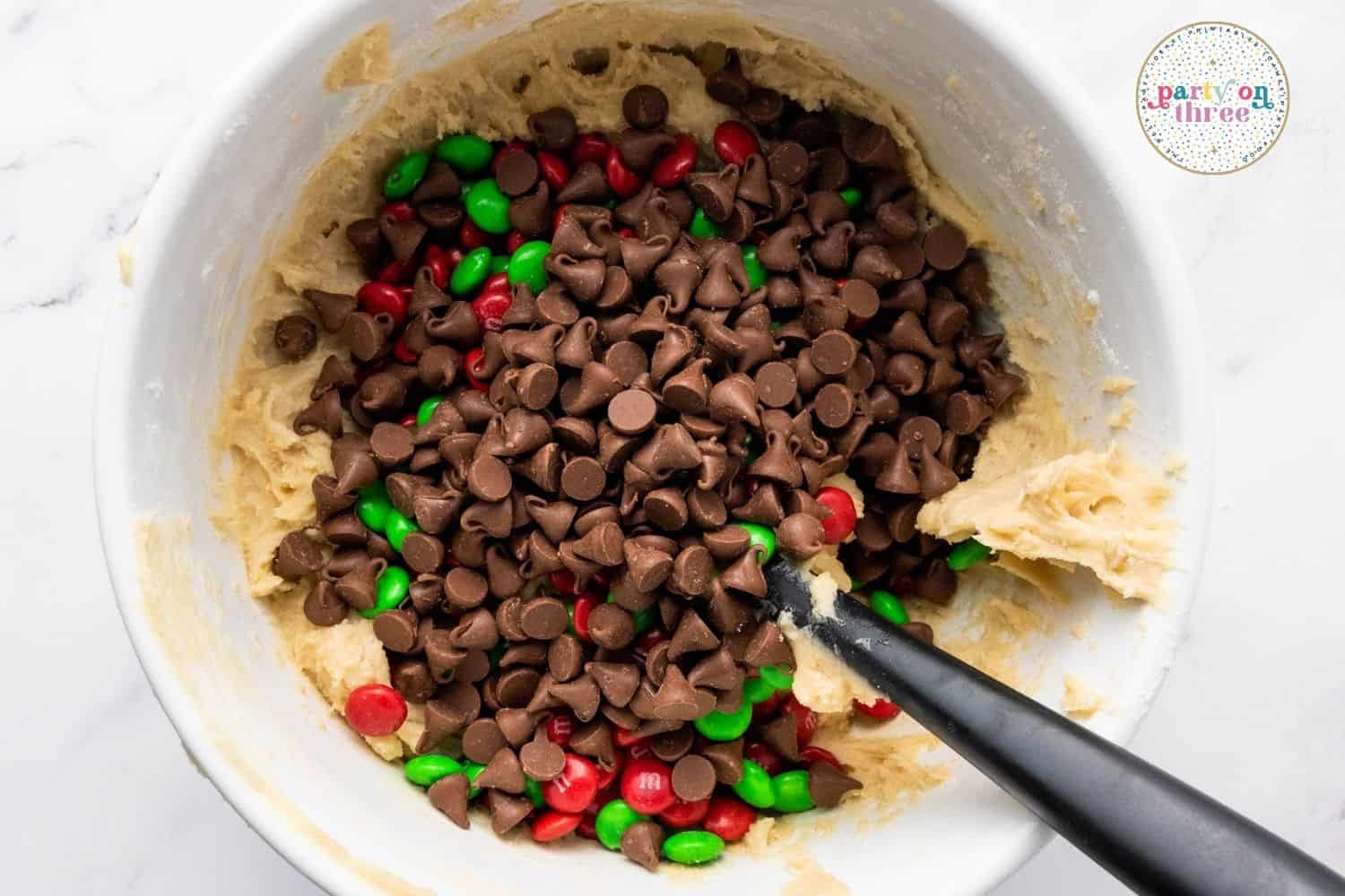 Chocolate chips and red and green M&Ms being mixed into cookie dough in a bowl
