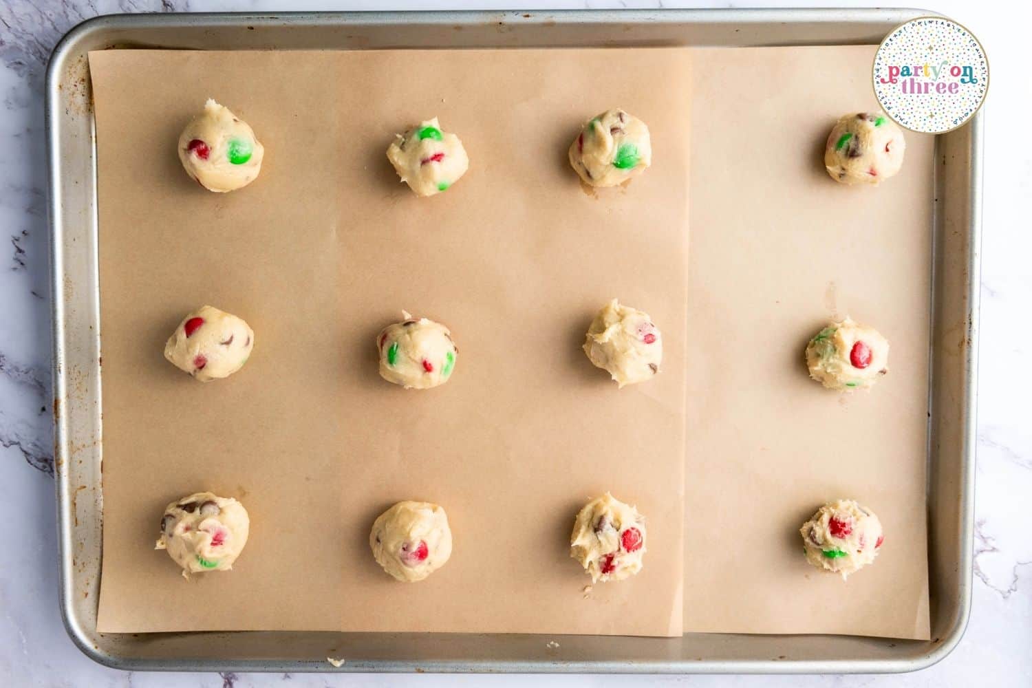 Dough balls with Christmas M&M's and chocolate chips arranged on a parchment-lined baking sheet