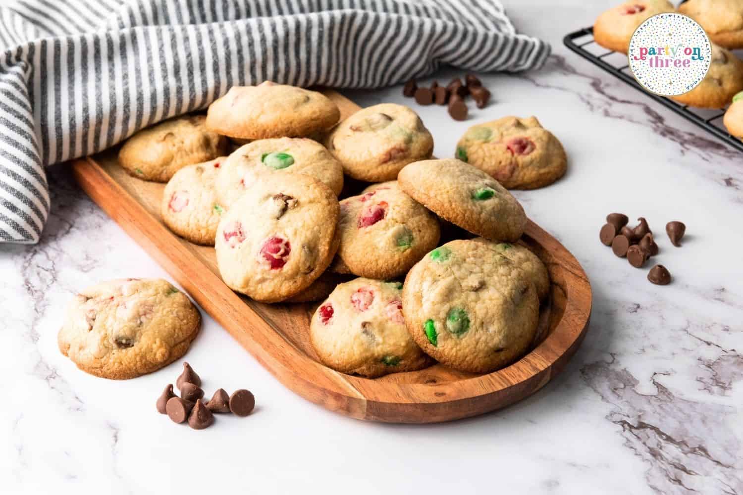 Baked Christmas M&M cookies piled on a wood board with chocolate chips scattered around