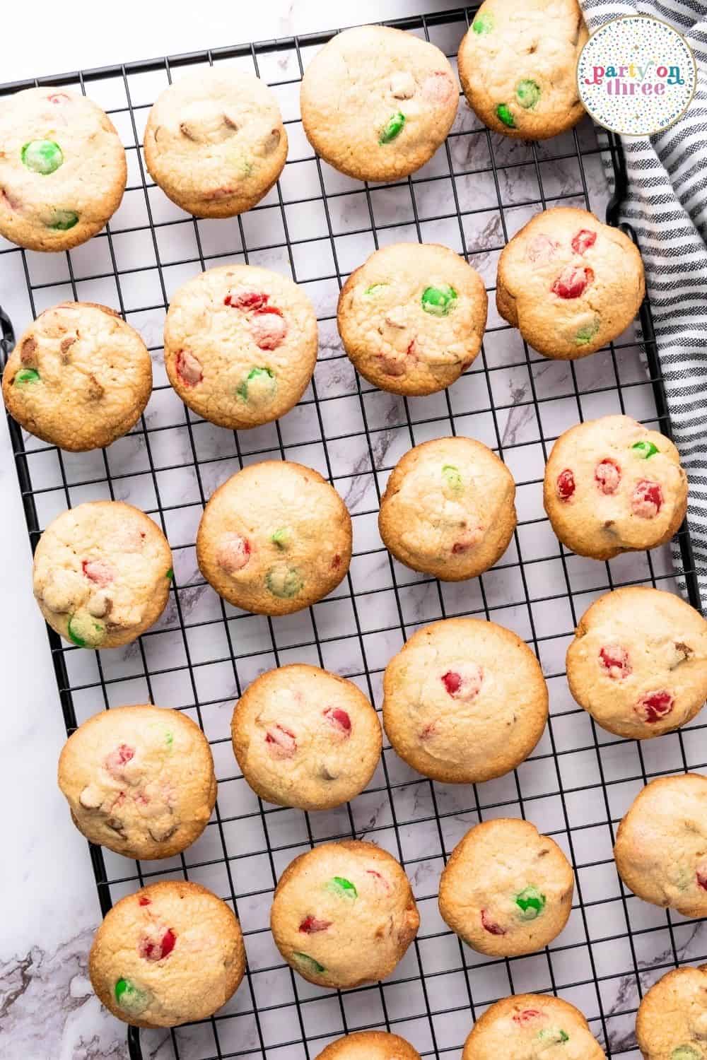 Rows of baked Christmas M&M cookies with red and green candies cooling on a black wire rack.