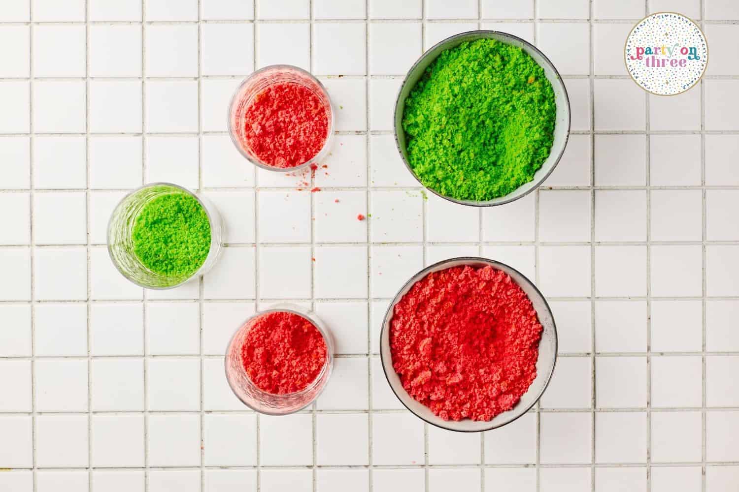Crumbled red velvet and green cake in small bowls on a white tiled surface