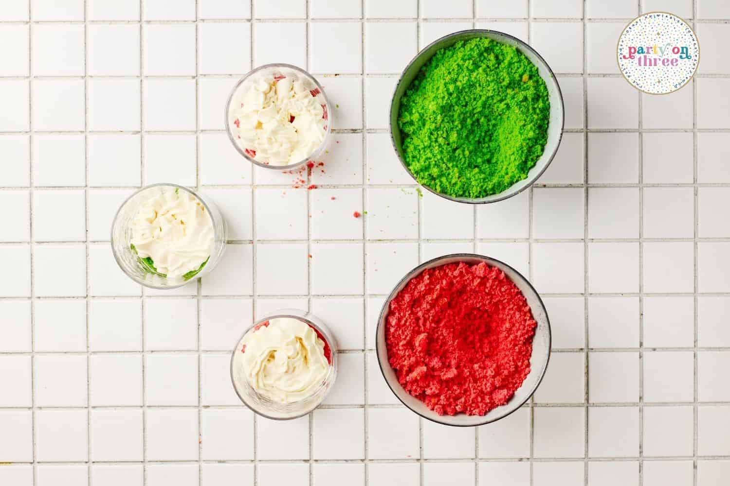 Bowls of crumbled red and green cake and small cups of whipped topping on a tiled surface