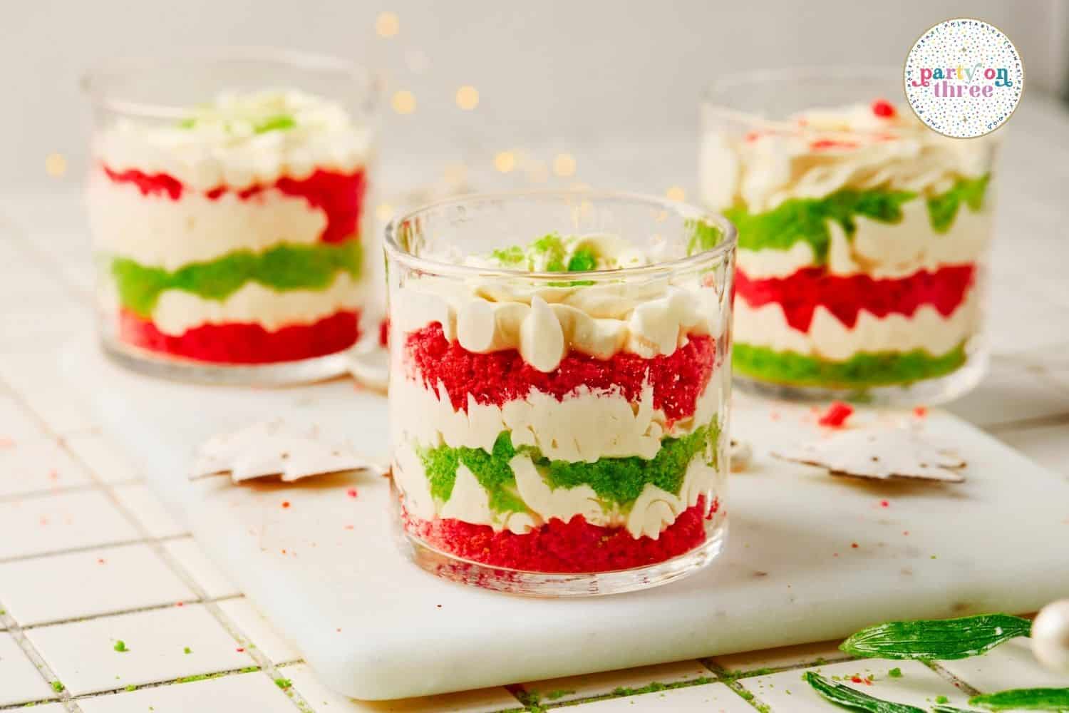 Three glass cups layered with red and green cake crumbs and creamy filling, topped with piped cream cheese mixture.