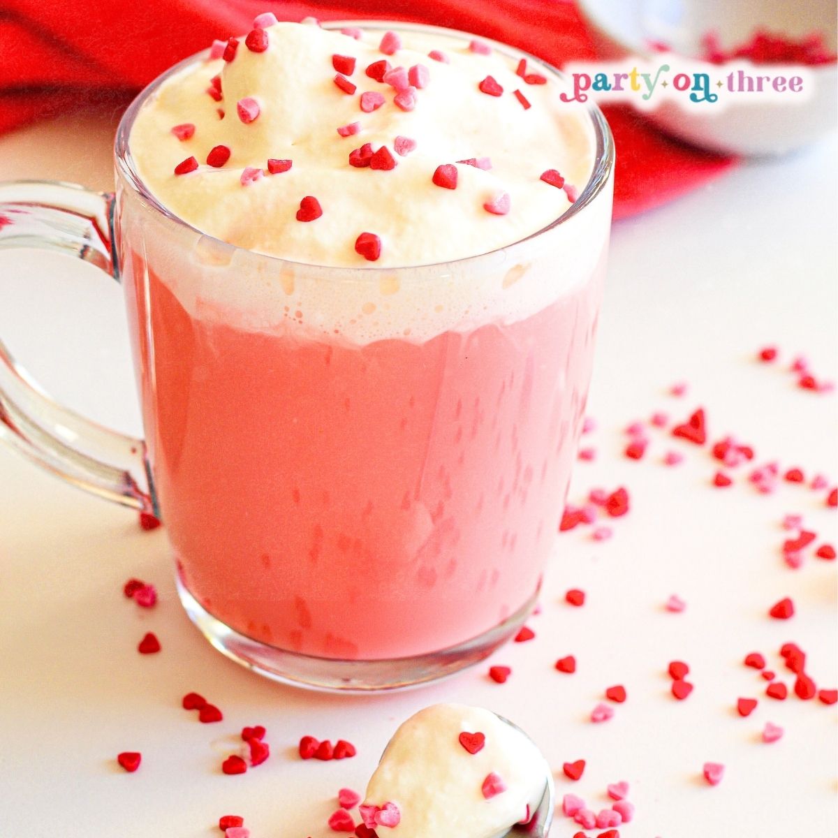 Pink hot chocolate in a glass mug topped with whipped cream and red and pink heart sprinkles