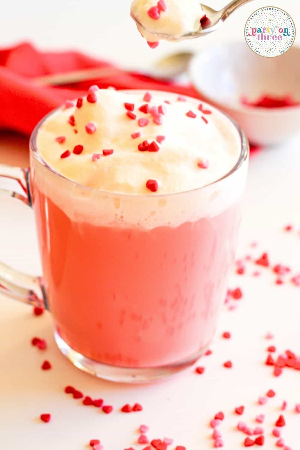 Glass mug of pink hot chocolate with whipped cream and heart sprinkles beside a red napkin