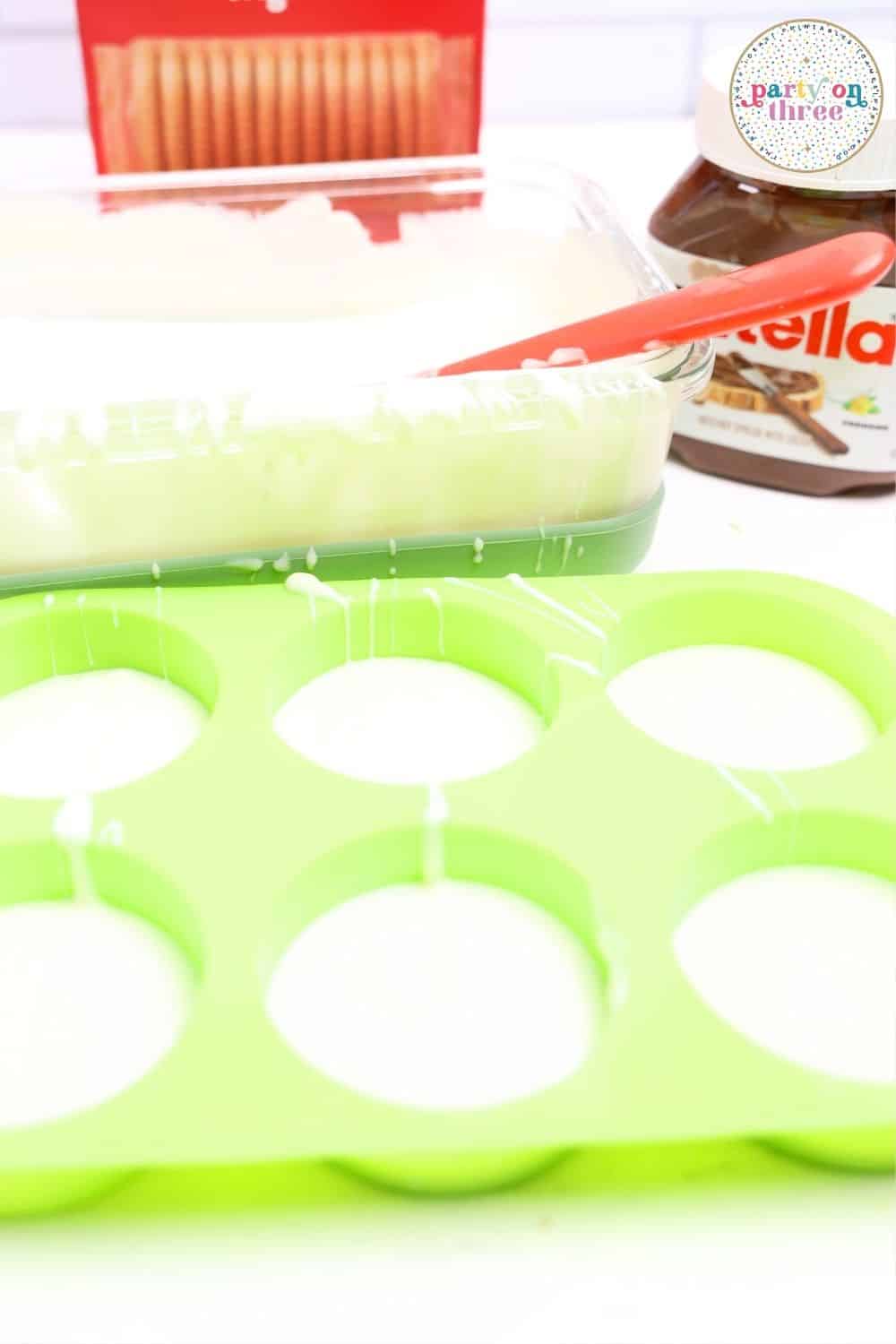 Melted white chocolate filling a green silicone round mold, with Ritz crackers and Nutella jar in background